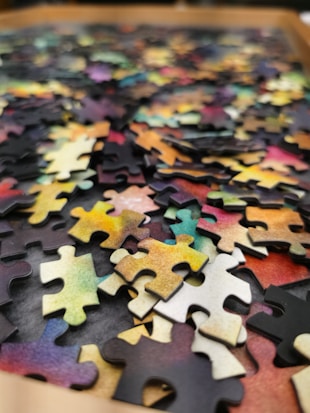 A collection of colorful jigsaw puzzle pieces scattered on a surface. The pieces display a variety of colors including shades of yellow, pink, green, and blue. The image focuses on the texture and complexity of the interlocking pieces, highlighting the randomness of their arrangement.