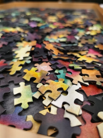 A collection of colorful jigsaw puzzle pieces scattered on a surface. The pieces display a variety of colors including shades of yellow, pink, green, and blue. The image focuses on the texture and complexity of the interlocking pieces, highlighting the randomness of their arrangement.