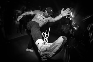 A monochrome image capturing a dynamic scene where a person is performing an energetic dance move, extending one leg towards the camera. The person is wearing a hat, a balaclava, a t-shirt, and sneakers. There are other people in the background with blurred effects, suggesting motion and a lively atmosphere.
