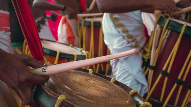 A group of individuals dressed in colorful traditional attire play large hand-crafted drums. The focus is on the intricate details of the drums, including their wooden surfaces, rope bindings, and the players’ precise hand movements with drumsticks.