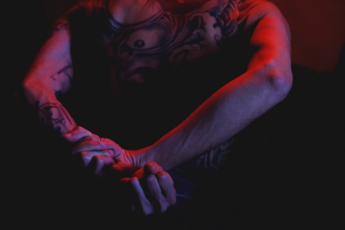 A person with detailed tattoos on their arm and chest is seen in a dark, moody setting, illuminated by red and blue lighting. The lighting emphasizes the contours of their muscles and tattoos, creating a dramatic effect.