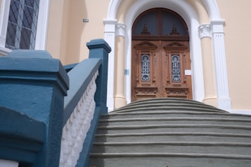 A grand entrance with a double wooden door featuring intricate designs and glass panels. The doors are flanked by ornate white columns and are at the top of a wide, curved staircase. The railing of the staircase is painted in teal and white, complementing the beige facade of the building.