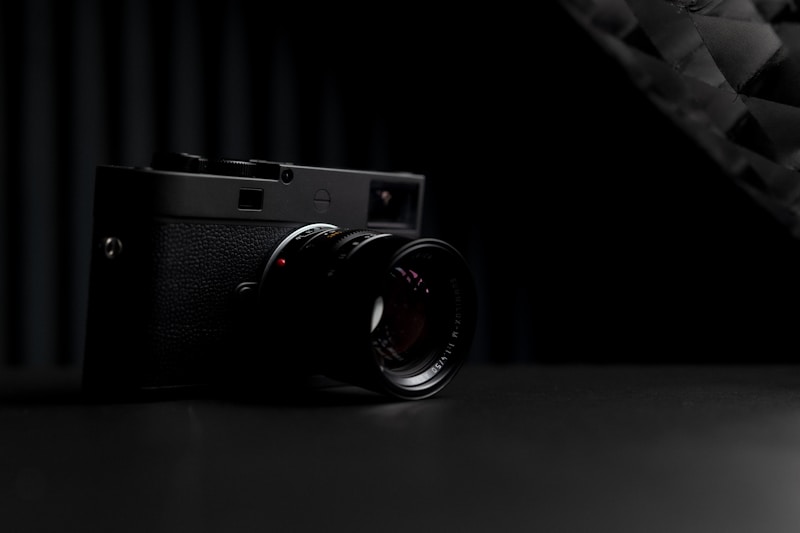 A black, vintage-style camera is positioned on a dark surface. The lighting is low, accentuating the camera's details and creating a dramatic shadow. The background appears to be dark, contributing to the overall mysterious and elegant atmosphere.