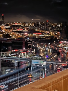 A bustling cityscape at night featuring a network of illuminated roadways and highways, densely packed buildings, and vibrant neon signs atop commercial complexes and skyscrapers. The urban environment is alive with the motion of vehicles and the glow of city lights reflecting off wet surfaces, indicating recent rainfall.