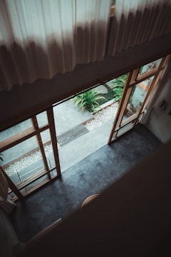 An aerial view from an upper level, showcasing a living area with large wooden-framed glass doors leading to an outdoor patio. The floor is smooth, likely concrete, with a contrast between indoor and outdoor elements. The patio area includes a row of white stones and potted greenery, providing a natural touch against the minimalistic design.