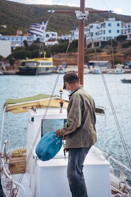A person stands on a boat holding a blue bag, facing away toward a marina with several boats docked. The background features hillside houses and a Greek flag flying on the boat.