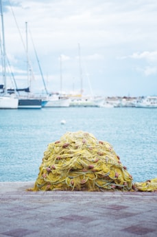 A large pile of yellow fishing nets sits on a stone dock by the water. Orange floats are interspersed throughout the tangled nets. Behind, several boats and yachts are moored in the marina, set against a cloudy blue sky.