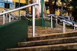 A series of outdoor stone steps with metal railings leading upward, flanked by green panels. Trees and a yellow building with windows are visible in the background, with several signs and logos displayed on the structure.