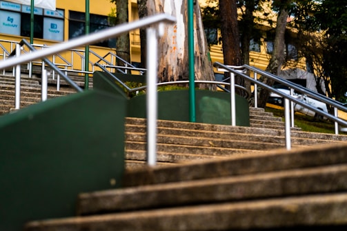 A series of outdoor stone steps with metal railings leading upward, flanked by green panels. Trees and a yellow building with windows are visible in the background, with several signs and logos displayed on the structure.