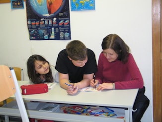 A woman and two children are sitting at a desk, engaging in a learning activity with books and pens. A large educational poster with diagrams and blue backgrounds is hanging on the wall behind them.