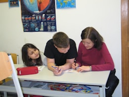 A woman and two children are sitting at a desk, engaging in a learning activity with books and pens. A large educational poster with diagrams and blue backgrounds is hanging on the wall behind them.