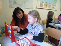 A young girl interacts with an abacus while seated at a desk, accompanied by a woman who appears to be assisting her. The setting suggests a classroom environment, with educational materials and a world map visible in the background. Other children are seated at their desks in the background.