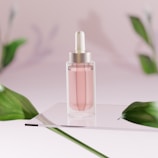 A clear glass dropper bottle containing a pink liquid is positioned on a reflective surface. The background is softly blurred with a pink hue and features green leaves that frame the scene, adding a natural element.