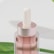 A close-up of a cosmetic bottle with a dropper lid. The bottle is partially filled with a pink liquid and has a silver metallic cap. The background is white with green leaves partially visible.