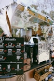 A mobile coffee cart adorned with white flowers and decorative grasses. It features a black menu board displaying various coffee options with prices, such as espresso, cappuccino, and cafe latte. Behind the board, there is a shiny coffee machine and various items neatly placed around it, including wicker baskets. The atmosphere appears relaxed and charming.