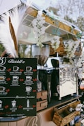 A mobile coffee cart adorned with white flowers and decorative grasses. It features a black menu board displaying various coffee options with prices, such as espresso, cappuccino, and cafe latte. Behind the board, there is a shiny coffee machine and various items neatly placed around it, including wicker baskets. The atmosphere appears relaxed and charming.