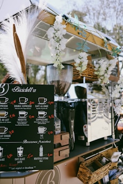 A mobile coffee cart adorned with white flowers and decorative grasses. It features a black menu board displaying various coffee options with prices, such as espresso, cappuccino, and cafe latte. Behind the board, there is a shiny coffee machine and various items neatly placed around it, including wicker baskets. The atmosphere appears relaxed and charming.