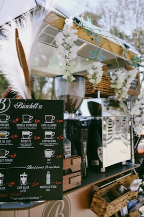 A mobile coffee cart adorned with white flowers and decorative grasses. It features a black menu board displaying various coffee options with prices, such as espresso, cappuccino, and cafe latte. Behind the board, there is a shiny coffee machine and various items neatly placed around it, including wicker baskets. The atmosphere appears relaxed and charming.
