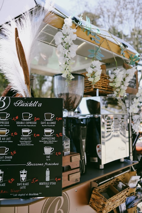 A mobile coffee cart adorned with white flowers and decorative grasses. It features a black menu board displaying various coffee options with prices, such as espresso, cappuccino, and cafe latte. Behind the board, there is a shiny coffee machine and various items neatly placed around it, including wicker baskets. The atmosphere appears relaxed and charming.