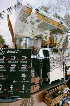 A mobile coffee cart adorned with white flowers and decorative grasses. It features a black menu board displaying various coffee options with prices, such as espresso, cappuccino, and cafe latte. Behind the board, there is a shiny coffee machine and various items neatly placed around it, including wicker baskets. The atmosphere appears relaxed and charming.