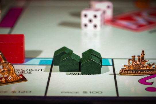 The image features a close-up view of a Monopoly game board. Visible are green houses, a red hotel, a pair of white dice with black dots, and gold game pieces including a battleship and a cannon. The game board sections are marked with property names and prices.