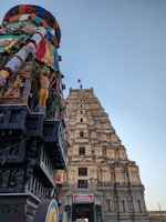 A richly decorated chariot adorned with colorful fabrics and intricate carvings stands beside a grand, ancient temple tower. The temple tower features multiple tiers embellished with sculptures. A red flag is visible on top of the tower, set against a clear blue sky.