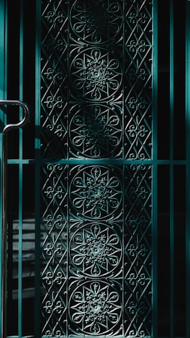 An ornate iron gate featuring intricate floral and geometric patterns. The design is symmetrical, with circular and diamond-shaped elements. The light casts shadows that enhance the depth and complexity of the patterns.