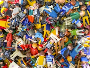 A scattered collection of various colorful toy figures, predominantly made of plastic. Many of the figures are wearing different uniforms and outfits, suggesting themes like police, sports, and casual attire. They are randomly piled on top of each other, creating a chaotic and vibrant scene.