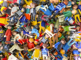 A scattered collection of various colorful toy figures, predominantly made of plastic. Many of the figures are wearing different uniforms and outfits, suggesting themes like police, sports, and casual attire. They are randomly piled on top of each other, creating a chaotic and vibrant scene.