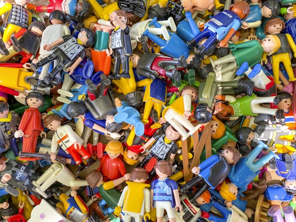 A scattered collection of various colorful toy figures, predominantly made of plastic. Many of the figures are wearing different uniforms and outfits, suggesting themes like police, sports, and casual attire. They are randomly piled on top of each other, creating a chaotic and vibrant scene.