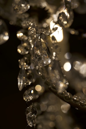 A close-up view of a crystal chandelier with intricate details. The image captures multiple faceted crystals reflecting light, creating a sparkling and elegant effect. The background is softly blurred, enhancing the focus on the central crystal elements.