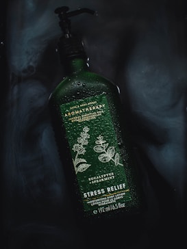 A green bottle with a pump dispenser labeled 'Aromatherapy Stress Relief'. The label features illustrations of eucalyptus and spearmint leaves. The bottle is covered in water droplets, resting on a dark surface with a smoky texture in the background.