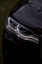 A close-up shot of a car's headlight. The headlight has a modern, angular design with bright LED lights illuminated. The surface of the car appears glossy, reflecting the sky and surroundings. The background is slightly blurred with hints of greenery, suggesting the car is parked outdoors.