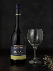 A bottle of Grand Tokaj Tokaji Amethyst wine labeled Hárslevelű 2021 stands next to an empty wine glass. The background is dark, enhancing the elegance of the scene. A small, delicate sprig of dried flowers rests near the base.