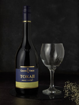 A bottle of Grand Tokaj Tokaji Amethyst wine labeled Hárslevelű 2021 stands next to an empty wine glass. The background is dark, enhancing the elegance of the scene. A small, delicate sprig of dried flowers rests near the base.