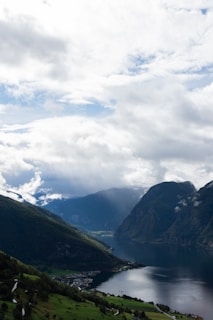 A serene landscape featuring a fjord surrounded by majestic mountains under a partly cloudy sky. Sunlight breaks through the clouds, casting soft rays onto the water and lush green hills. A small village is nestled in the valley, with winding roads leading to it.