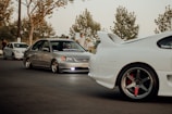 A group of modified cars, including a silver Honda Civic and a white sports car with a prominent rear spoiler, parked on a street. The scene includes people standing nearby under trees with sparse leaves, suggesting an autumn setting. The lighting is warm and subdued, giving a laid-back feel.