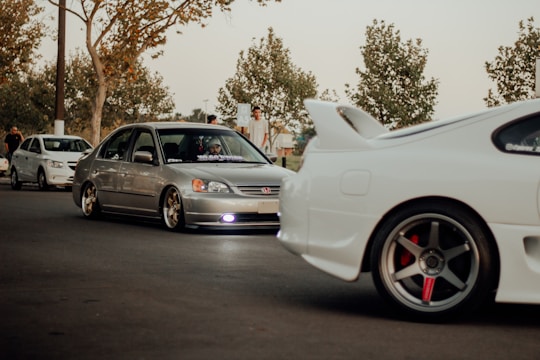 A group of modified cars, including a silver Honda Civic and a white sports car with a prominent rear spoiler, parked on a street. The scene includes people standing nearby under trees with sparse leaves, suggesting an autumn setting. The lighting is warm and subdued, giving a laid-back feel.