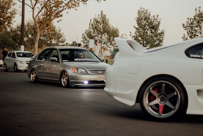A group of modified cars, including a silver Honda Civic and a white sports car with a prominent rear spoiler, parked on a street. The scene includes people standing nearby under trees with sparse leaves, suggesting an autumn setting. The lighting is warm and subdued, giving a laid-back feel.