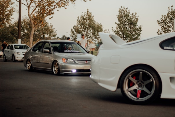 A group of modified cars, including a silver Honda Civic and a white sports car with a prominent rear spoiler, parked on a street. The scene includes people standing nearby under trees with sparse leaves, suggesting an autumn setting. The lighting is warm and subdued, giving a laid-back feel.