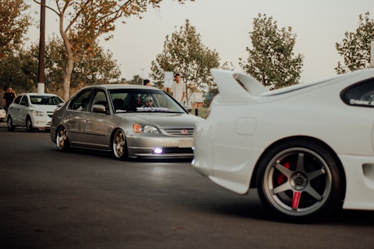 A group of modified cars, including a silver Honda Civic and a white sports car with a prominent rear spoiler, parked on a street. The scene includes people standing nearby under trees with sparse leaves, suggesting an autumn setting. The lighting is warm and subdued, giving a laid-back feel.