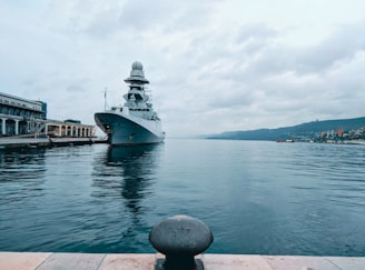 A large naval ship is docked at a port, with a sizable structure building nearby. The water is calm, and there are hills in the background under an overcast sky.