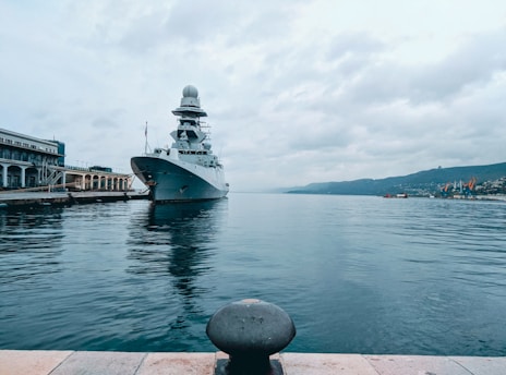 A large naval ship is docked at a port, with a sizable structure building nearby. The water is calm, and there are hills in the background under an overcast sky.