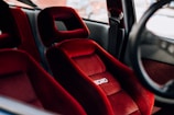 Two vibrant red Recaro car seats with a plush texture, featuring a checkered pattern, are positioned inside a vehicle. The seats are designed for comfort and sportiness, evident by the ergonomic shape. The interior of the car is partially visible, with a seatbelt and part of the steering wheel seen in the background.