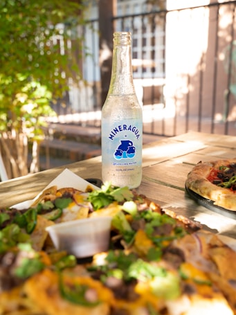 A clear bottle of sparkling water branded Mineragua is placed on a wooden table. Surrounding the bottle are plates of food, including a pizza and a dish garnished with various toppings and fresh greens. Sunlight filters through the surrounding space, casting soft shadows around the scene.