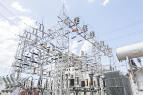 A complex network of power lines and electrical equipment, including transformers and insulators, spans across a substation structure. Numerous high-voltage cables connect various components, set against a backdrop of a partly cloudy sky.
