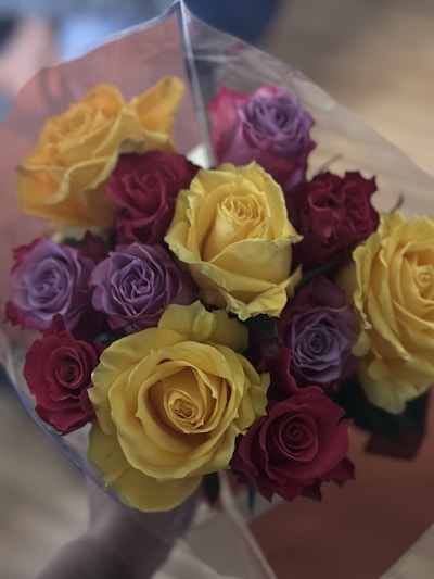 A bouquet of vibrant flowers featuring yellow, red, and purple roses. The roses are fresh, with velvety petals and are arranged tightly together in a clear wrapping.