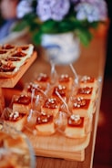 A variety of desserts arranged neatly on wooden trays, featuring caramel-topped squares with small white decorations. In the background, a blurred floral arrangement in a decorative vase adds to the setting's elegance.