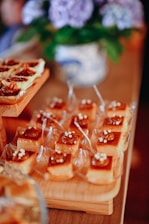A variety of desserts arranged neatly on wooden trays, featuring caramel-topped squares with small white decorations. In the background, a blurred floral arrangement in a decorative vase adds to the setting's elegance.
