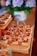 A variety of desserts arranged neatly on wooden trays, featuring caramel-topped squares with small white decorations. In the background, a blurred floral arrangement in a decorative vase adds to the setting's elegance.